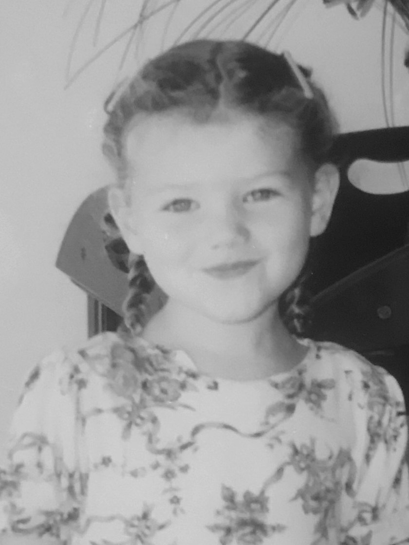 A childhood photo: a young girl with braided pigtails and a floral dress, smiling.